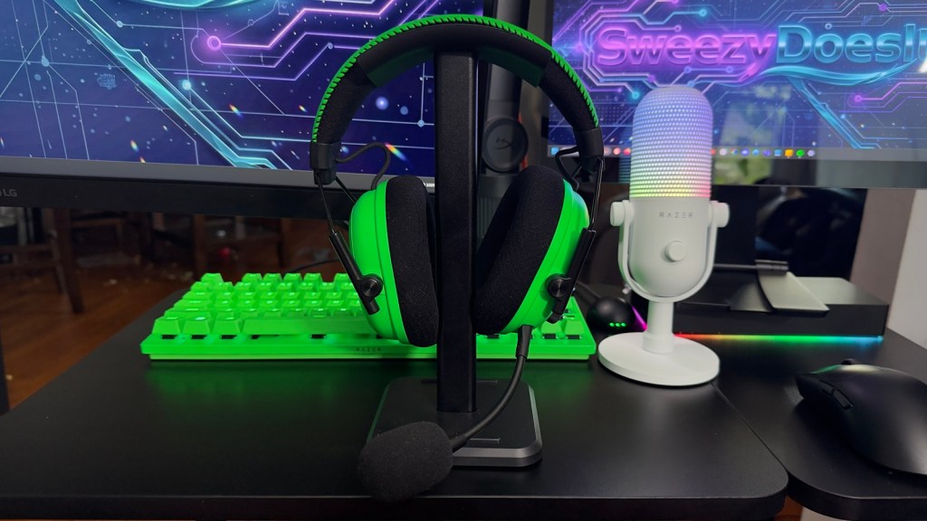 A gaming headset in green resting on a stand, alongside a green mechanical keyboard and a white microphone with RGB lighting, set against a computer monitor displaying a colorful digital design.