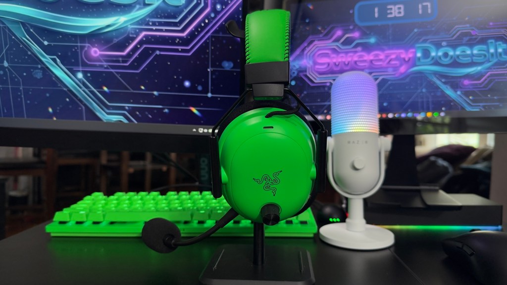 A green gaming headset on a stand, with a matching green keyboard in the background. There is a colorful microphone beside the headset, and a computer monitor displaying the text 'Sweezy Does It' with a digital clock.