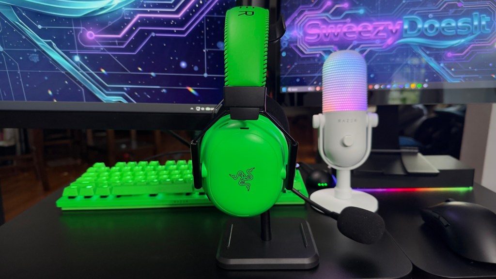 A vibrant green gaming headset displayed on a stand, in front of a computer monitor with a digital background featuring the text 'Sweezy Does It'. Nearby, there is a white microphone with colorful LED lighting and a green keyboard.
