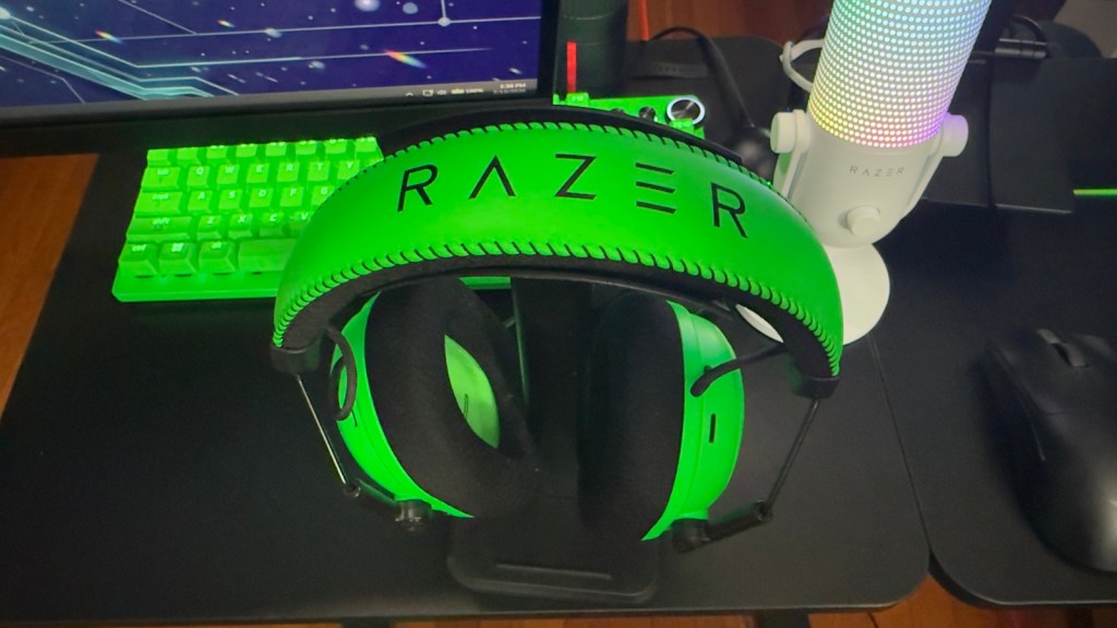Green Razer gaming headset resting on a stand, with a gaming keyboard and a colorful microphone in the background.
